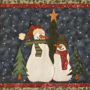 Może przedstawiać: Wiszące ozdoby ścienne w postaci dwóch bałwanów stojących obok zielonej choinki. Bałwany mają na sobie szaliki i czapki. Tło jest ciemnoniebieskie z białymi płatkami śniegu.