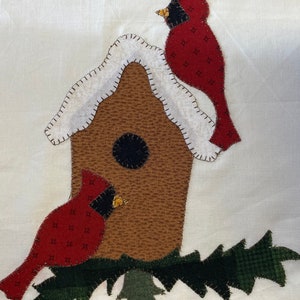 May include: A brown birdhouse with a white snowy roof, two red cardinals, and green foliage.