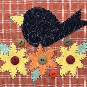 May include: A black bird with a yellow beak is perched on top of three orange and yellow flowers with green leaves. The flowers have brown buttons in the center. The background is a red and white checkered fabric.