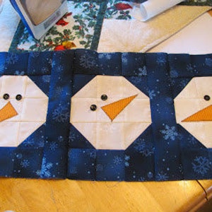 May include: A blue and white fabric panel with three snowman faces. Each snowman face has two black buttons for eyes and an orange felt nose.