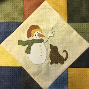 May include: A white snowman with a brown carrot nose and a green scarf is holding a stick with a brown dog sitting next to it. The snowman and dog are on a yellow background with a green, brown, blue and yellow diamond pattern.