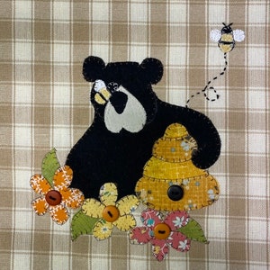 May include: A black bear with a beehive behind it, surrounded by colorful flowers with buttons in the center. A bee flies above the bear.
