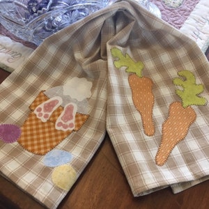 May include: A beige and white checkered tea towel with a bunny rabbit and carrots appliqued on it. The bunny is in a basket with pink ears and feet. The carrots are orange and green.