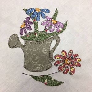 Puede incluir: Una regadera gris con un patrón floral está llena de un ramo de flores coloridas. Las flores son moradas, azules y rosas con centros amarillos. La regadera tiene un pico y un asa. Las flores están rodeadas de hojas verdes.