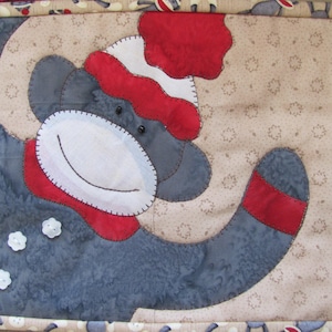 Può includere: Una scimmia grigia e rossa con un cappello bianco e una sciarpa a righe rosse e bianche. La scimmia sorride e ha un bottone bianco sul petto. La scimmia è su uno sfondo beige con un motivo floreale.