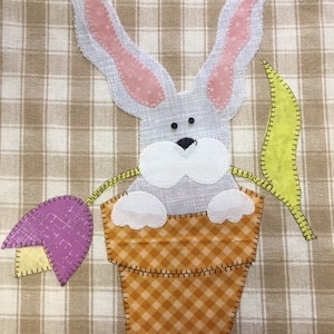 May include: A gray bunny with pink ears is sitting in a brown and orange checkered pot. The bunny is peeking out of the pot and has a white nose and black eyes. There is a purple flower with a yellow stem next to the pot.