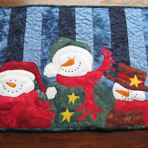 Könnte beinhalten: Ein gesteppter Tischläufer mit Winterthema mit drei Schneemännern mit roten Schals und Mützen. Die Schneemänner befinden sich auf einem blau-grünen gestreiften Hintergrund mit Schneeflocken.