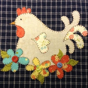 May include: A white fabric chicken with a red comb and a floral patterned tail, surrounded by colourful fabric flowers with buttons in the centre. The chicken and flowers are appliquéd on a dark blue checked fabric.