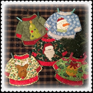 May include: Five handmade Christmas decorations shaped like jumpers with different festive designs. The decorations are made of fabric and have a red, green and white colour scheme. The decorations are hanging from a wire with clothes pegs.