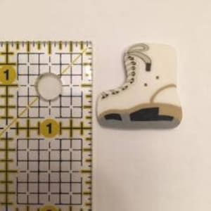 May include: A small, off-white ice skate-shaped embellishment with brown and gray accents. The embellishment is approximately 1 inch in height and width, with a black sole and gray trim. The skate has a lace-up design.
