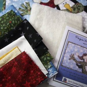 May include: A set of fabric scraps in various colors and patterns, including green, black, white, red, and yellow. The fabric scraps are arranged in a stack, with a white piece of batting on top. A printed pattern for a penguin mug rug is also included, with the text "Penguin Mug Rug #307 Quilt Doodle Designs".
