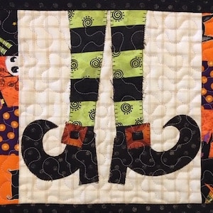 May include: A quilted Halloween-themed coaster featuring a pair of black witch's boots with green and black striped stockings. The boots have pointed toes and buckles. The background is a patchwork of various Halloween-themed fabrics.