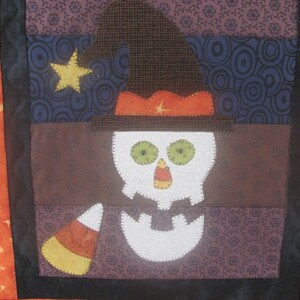 May include: A patchwork quilt block featuring a white skull wearing a witch's hat with a brown brim and orange band. The skull has green eyes and a candy corn nose. The background is a mix of brown, purple, and blue fabrics with various patterns.