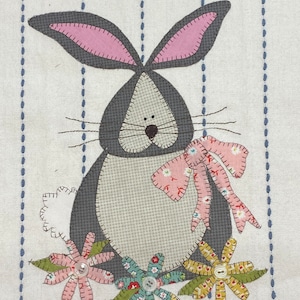 May include: A gray bunny with pink ears and a bow is surrounded by colorful flowers. The bunny is stitched onto a white fabric with blue stripes.