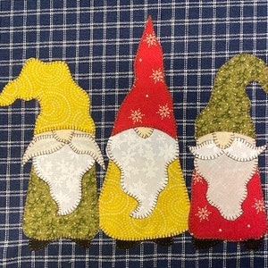 May include: Three gnomes with different colored hats and patterned fabric. The gnome on the left has a yellow hat, the middle gnome has a red hat, and the gnome on the right has a green hat.