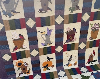 Könnte beinhalten: Eine lila und weiße Patchwork-Decke mit verschiedenen Motiven von Vögeln, Blumen und Häusern. Die Decke ist aus verschiedenen Stoffen und Texturen gefertigt und erzeugt ein optisch ansprechendes und komplexes Design.