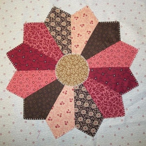 May include: A quilt block with a starburst design in shades of red, brown, and beige. The center of the block is a circle with a floral pattern.