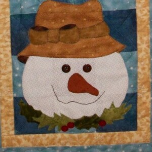May include: A snowman wearing a brown hat and a green holly wreath around its neck. The snowman has a red nose and black eyes. The background is a blue and white quilted fabric.