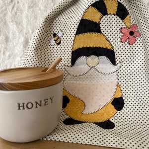 May include: A cream-colored ceramic honey pot with a wooden lid and spoon sits next to a decorative kitchen towel. The towel features a gnome design with a yellow and black striped hat, a pink flower, and a bee.