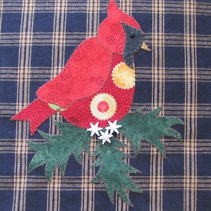 Może przedstawiać: Czerwony kardynał z czarnymi oczami i żółtym dziobem siedzący na zielonej, liściastej gałęzi z białymi płatkami śniegu. Ptak znajduje się na niebieskim i brązowym wzorze w kratę.