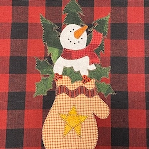 May include: A snowman wearing a red scarf and a green holly wreath is nestled inside a red and white checkered mitten with a yellow star. The mitten is on a red and black buffalo plaid background.
