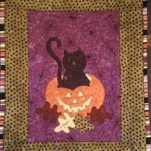 Puede incluir: Un gato negro se sienta dentro de una calabaza tallada con una sonrisa de dientes. La calabaza está rodeada de hojas verdes y el fondo es una tela morada con un patrón de telaraña.