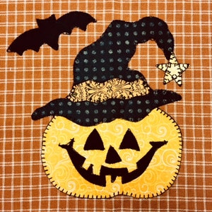 May include: A yellow pumpkin with a black witch hat and a black bat flying above. The pumpkin has a carved face with a wide smile and three triangles for eyes. The hat has a black and white polka dot pattern and a gold trim.