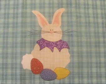 May include: A white bunny with purple and floral fabric around its neck, holding three Easter eggs. The eggs are red, blue, and yellow. The bunny is on a blue and white plaid background.