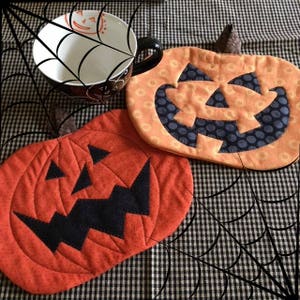 May include: Two orange and black pumpkin-shaped pot holders with stitched details. The pot holders are on a black and white checkered background.