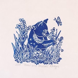 May include: Blue linocut print of a bear cub sniffing a flower with a butterfly flying above. The print is titled "Bear and Bluebells" and signed "Daynz".