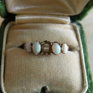 Puede incluir: Un anillo de oro con cinco opales blancos ovalados engastados en fila. El anillo está en una caja de anillo de terciopelo verde.