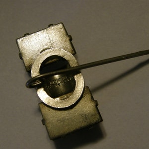 Puede incluir: Un pin de metal plateado con un dise&ntilde;o cuadrado y un cierre de pin en la parte posterior. El pin tiene una abertura circular con un bucle de metal y el texto "MADE IN U.S.A." es visible.