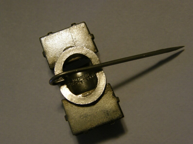 Puede incluir: Un pin de metal plateado con un dise&ntilde;o cuadrado y un cierre de pin en la parte posterior. El pin tiene una abertura circular con un bucle de metal y el texto "MADE IN U.S.A." es visible.
