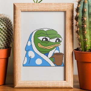 May include: A framed cross-stitch art piece featuring a green frog character wrapped in a blue blanket with white polka dots, holding a brown mug. The frame is surrounded by potted cacti, a camera, and other plants.
