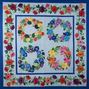 May include: A blue quilted fabric wall hanging with a floral design. The design features four wreaths of different flowers, including red, pink, purple, and white flowers. The wreaths are surrounded by a border of green leaves and flowers.