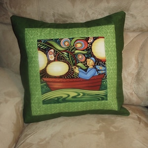 May include: A green pillow with a colorful illustration of a person in a boat surrounded by flowers and lights. The pillow has a green border with a decorative pattern.