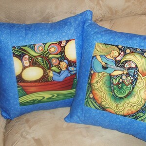 Puede incluir: Dos fundas de coj&iacute;n azules con dise&ntilde;os coloridos y caprichosos. Una funda presenta una sirena tocando la guitarra, la otra presenta a una persona en un bote con un &aacute;rbol y fruta.