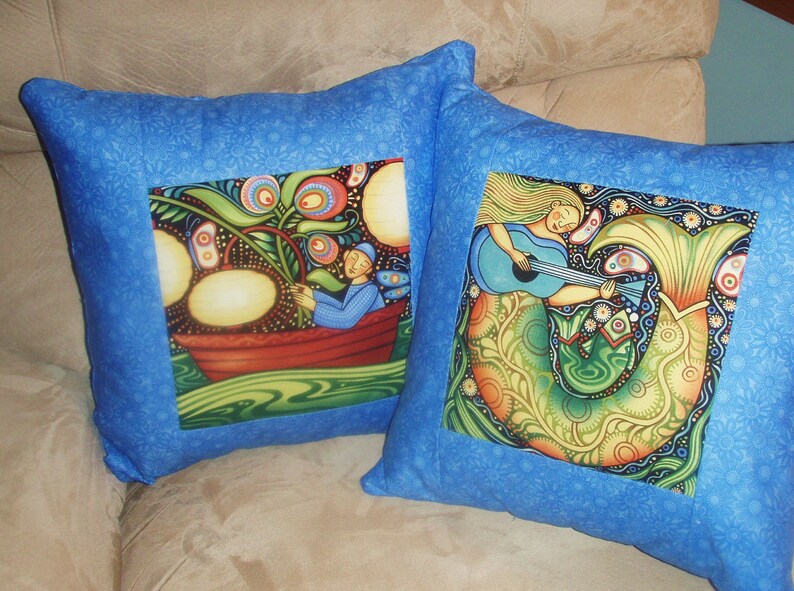 Puede incluir: Dos fundas de coj&iacute;n azules con dise&ntilde;os coloridos y caprichosos. Una funda presenta una sirena tocando la guitarra, la otra presenta a una persona en un bote con un &aacute;rbol y fruta.