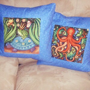 Puede incluir: Dos fundas de coj&iacute;n azules con dise&ntilde;os bordados coloridos. Una funda de coj&iacute;n presenta una rana verde con una corona montada en una tortuga azul. La otra funda de coj&iacute;n presenta un pulpo rojo con algas verdes y peces.