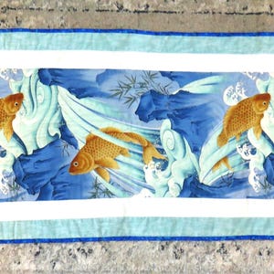May include: Rectangular quilted table runner featuring a central panel with a blue and white water scene and three orange goldfish. The runner is bordered by white and light blue fabric.