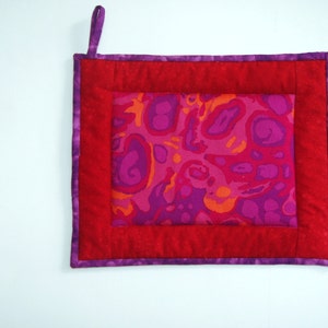 May include: A red and purple quilted hot pad with a pink and orange abstract pattern in the center. The hot pad has a loop for hanging.