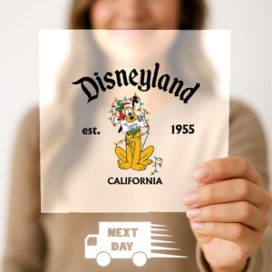 May include: A clear, ready-to-press DTF transfer featuring the Disneyland logo with Pluto. The design includes the text "Disneyland est. 1955 California" and a graphic of a truck with the words "Next Day".