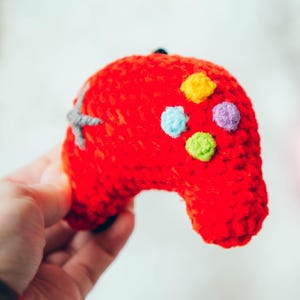 Puede incluir: Un mando de videojuego rojo tejido a mano con detalles de botones coloridos. El mando tiene una cruceta gris y botones coloridos. El fondo incluye ovillos de lana rosa y azul.