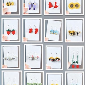 Può includere: Una collezione di orecchini all'uncinetto fatti a mano in vari design. Gli orecchini includono un'ape, uccelli, cappelli di Babbo Natale, girasoli, fragole, cuori, pulcini, orsacchiotti, conigli, gatti, bambole, alieni verdi e pulcini gialli.