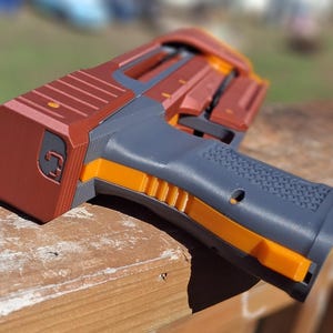 Puede incluir: Una pistola de juguete impresa en 3D, de aspecto futurista, en tonos óxido, gris y naranja. La empuñadura es texturizada y el diseño presenta una serie de líneas paralelas y formas geométricas. La pistola de juguete está sobre una superficie de madera.
