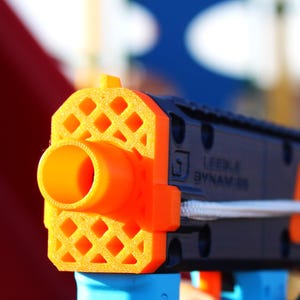 May include: A black and orange toy gun with a white string attached to the barrel. The gun has a waffle-like design on the barrel and the text "LEADER BYNAMIC" is printed on the side.