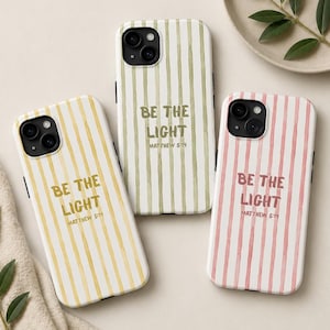 May include: Three smartphone cases featuring vertical watercolor stripes in yellow, olive green, and dusty pink. Each case displays the text BE THE LIGHT and MATTHEW 5:14 in a centered, sans-serif font. The cases have black camera lens cutouts.