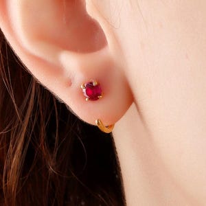 Könnte beinhalten: Nahaufnahme eines Ohrs mit einem kleinen roten Edelstein-Ohrring und einem goldenen Creolen-Ohrring.