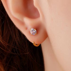 May include: A close-up of an ear with a small, round, gold hoop earring and a stud earring. The stud earring features a clear, round gemstone. Reddish-brown hair is visible in the background.