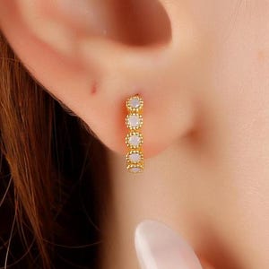 May include: A gold-colored earring with a series of small, round, opalescent stones. The earring is a small hoop style, worn in the ear. The stones are evenly spaced along the curve of the earring, creating a delicate and elegant look.